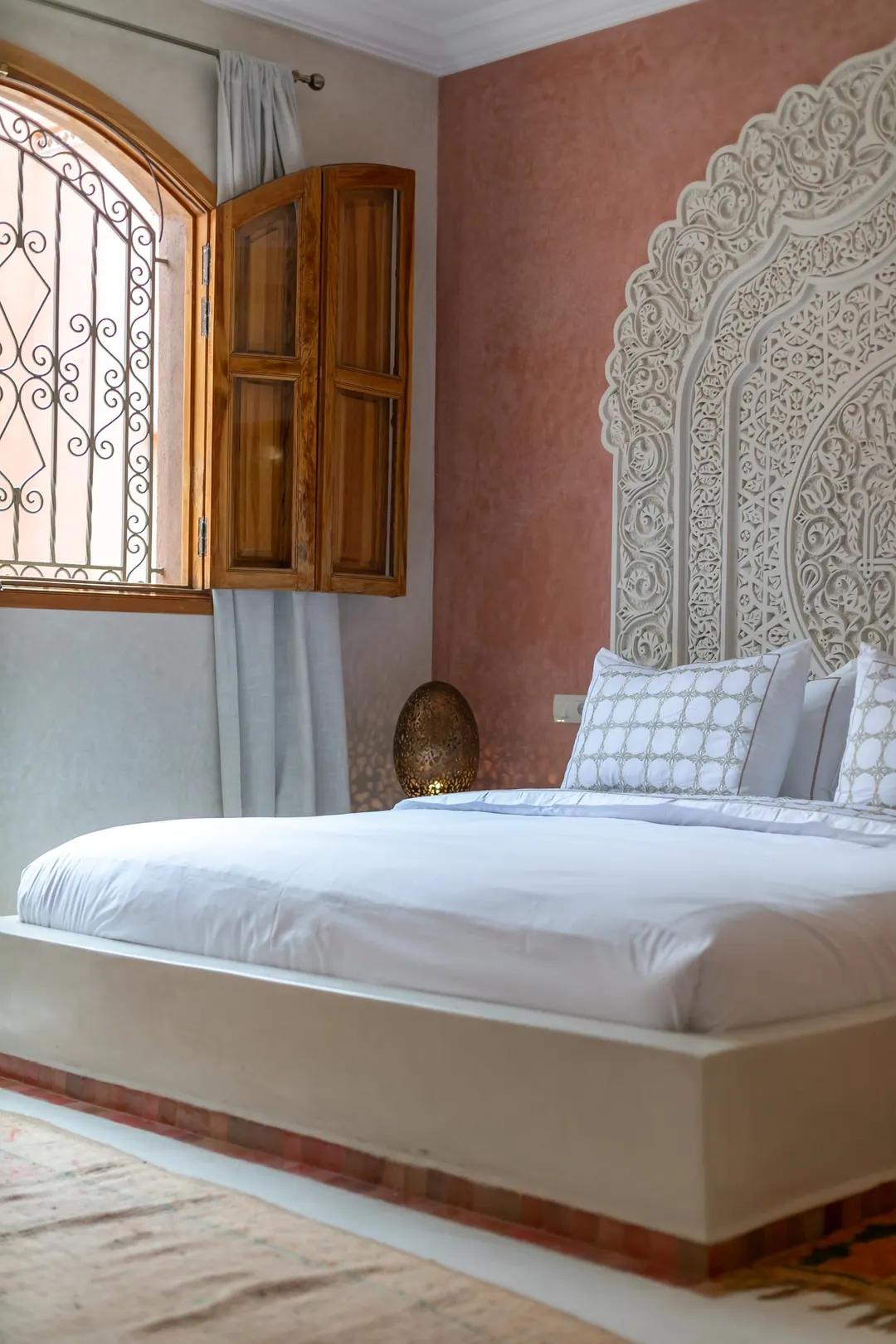 Terracotta room: carved white headboard on a rose-clay wall, cedar shutters and Berber rugs